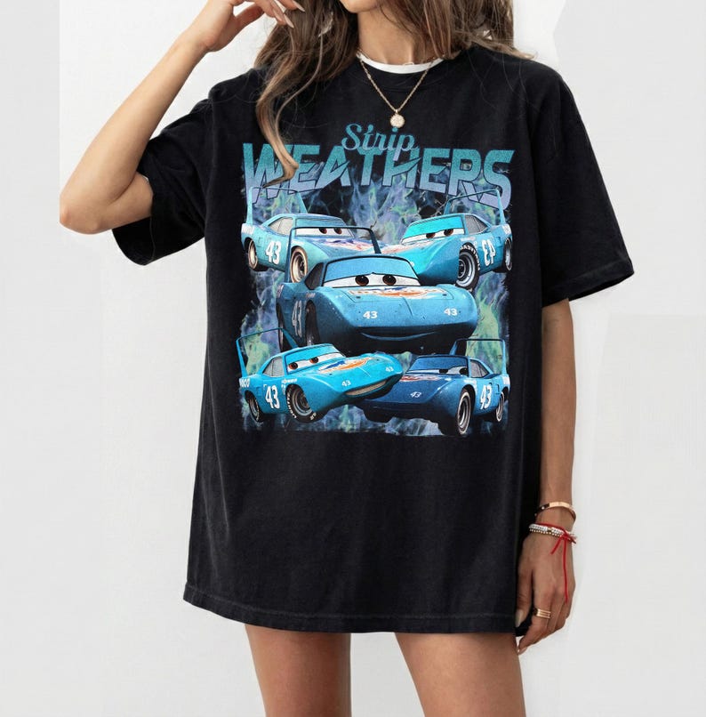 May include: Black t-shirt featuring a graphic of blue race cars with the number 43. The text "Strip Weathers" is above the cars. The shirt has a relaxed fit and short sleeves.