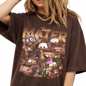 Puede incluir: Camiseta marr&oacute;n con un gr&aacute;fico del personaje Mater de la pel&iacute;cula Cars. El gr&aacute;fico incluye el texto "Tow Mater" en letras doradas. La camiseta tiene un aspecto vintage y desgastado.