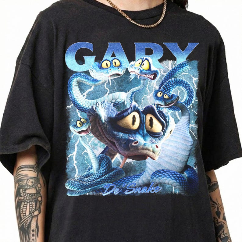 Pode incluir: Camiseta preta com um gr&aacute;fico de cobras azuis de desenho animado com olhos amarelos e o texto "GARY De Snake" em azul. As cobras est&atilde;o em um fundo de raios azuis.