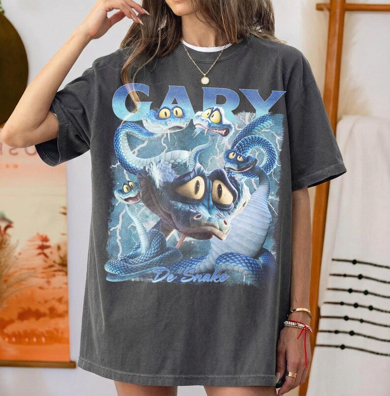 Pode incluir: Camiseta cinza escuro com uma ilustra&ccedil;&atilde;o de desenho animado de uma cobra azul com v&aacute;rias cabe&ccedil;as e o texto "GARY De Snake" em gradiente azul. O design inclui raios.