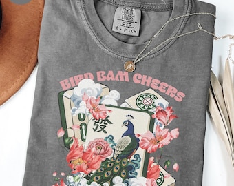 May include: A charcoal gray t-shirt featuring a graphic print of mahjong tiles, a peacock, and pink flowers. The text BIRD BAM CHEERS is printed in pink above the tiles. The shirt has a crew neckline and a Comfort Colors brand label inside.