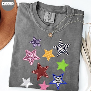 以下が含まれることがあります： チャコールグレーのTシャツで、ピンクのスパンコールの星、白黒の渦巻き模様の星、黄色の水玉模様の星、赤いステッチの星、ライムグリーンの星、青と白のゼブラ柄の星など、さまざまなカラフルな星のグラフィックがプリントされています。