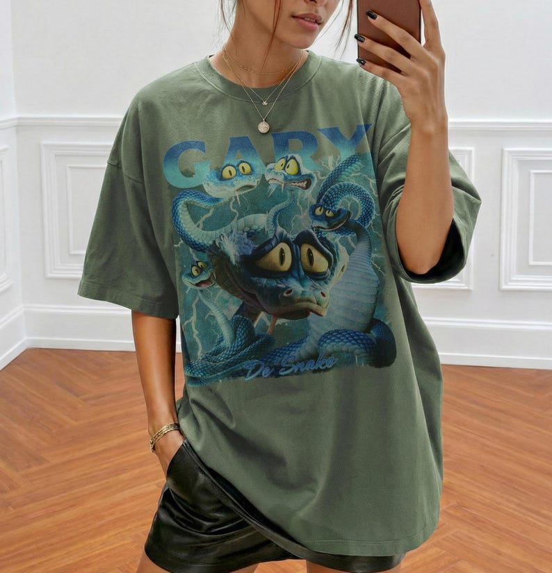 Pode incluir: Camiseta verde-oliva com um desenho de cobra de desenho animado. O desenho apresenta o texto "Gary De Snake" em azul, juntamente com v&aacute;rias cobras azuis com olhos amarelos e um fundo de rel&acirc;mpagos. A camiseta tem mangas curtas.