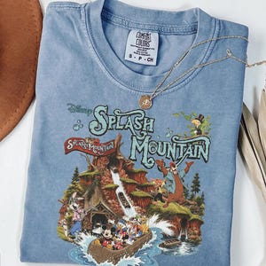 May include: A blue short-sleeve t-shirt featuring a graphic print of the Disney Splash Mountain attraction. The design includes characters like Mickey Mouse, Minnie Mouse, and Donald Duck in a log flume, with the text Splash Mountain printed above.