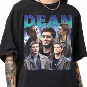 May include: Black t-shirt featuring a collage of images of a man in a leather jacket. The word "DEAN" is printed in blue and teal above the name "Winchester". The design has a vintage, distressed look.