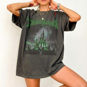 May include: Dark gray t-shirt with the word "Disneyland" in green, stylized text. The shirt features a green and black graphic of a castle with fireworks. The shirt has short sleeves and a relaxed fit.
