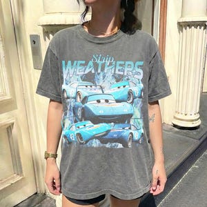 May include: A gray t-shirt featuring a graphic of blue race cars with the text "WEATHERS" in a light blue font. The cars have the number "43" on them. The shirt has a vintage, worn look.