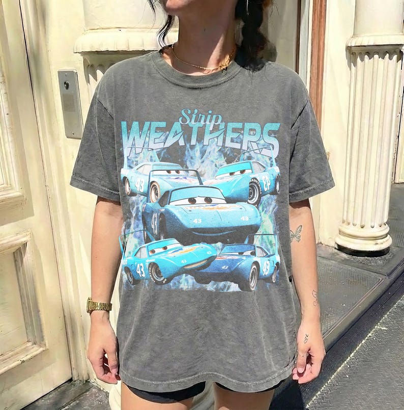 May include: A gray t-shirt featuring a graphic of blue race cars with the text "WEATHERS" in a light blue font. The cars have the number "43" on them. The shirt has a vintage, worn look.