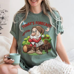 Op de afbeelding: Een saliegroen t-shirt met een afbeelding van een norse dwerg met een bruine muts, rood shirt en beige broek. De tekst luidt "TODAY'S FORECAST: Grumpy morning with a partially Grumpy afternoon" en "DAILY GEM" op een krant die de dwerg vasthoudt.