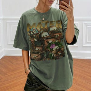 Puede incluir: Camiseta verde oliva de gran tama&ntilde;o con un gr&aacute;fico del personaje Tow Mater de la pel&iacute;cula Cars. El gr&aacute;fico incluye el texto "Tow Mater" en letras doradas y varios personajes de coches de dibujos animados. La camiseta tiene mangas cortas y un corte holgado.