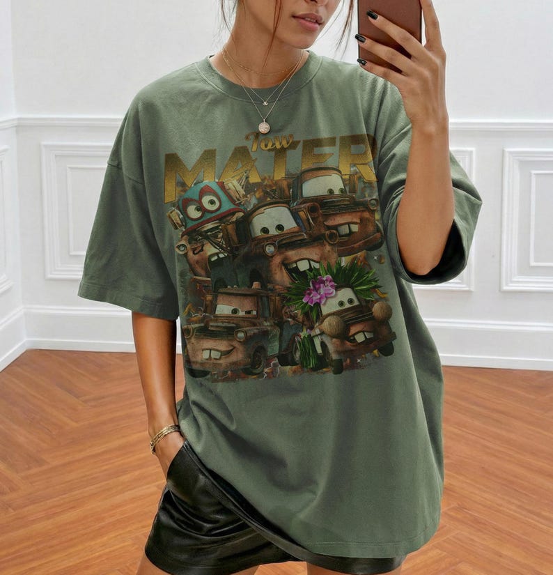 Puede incluir: Camiseta verde oliva de gran tama&ntilde;o con un gr&aacute;fico del personaje Tow Mater de la pel&iacute;cula Cars. El gr&aacute;fico incluye el texto "Tow Mater" en letras doradas y varios personajes de coches de dibujos animados. La camiseta tiene mangas cortas y un corte holgado.