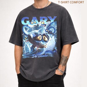Pode incluir: T-shirt cinzenta escura com um gr&aacute;fico de uma criatura azul semelhante a uma cobra com olhos grandes e raios. A palavra "GARY" est&aacute; no topo em letras azuis, e o texto "Do Snake" est&aacute; na parte inferior. A camisa est&aacute; rotulada como "PEPPER T-SHIRT COMFORT".