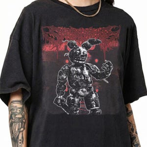 May include: Black t-shirt featuring a graphic of a robotic rabbit character with the word "SPRINGTRAP" in red above it. The design has a distressed, vintage look. The shirt is made of a soft material.