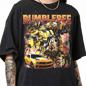 May include: Black t-shirt featuring the word "BUMBLEBEE" in red and yellow block letters. The graphic includes yellow robots, a yellow sports car, and a red and orange background.