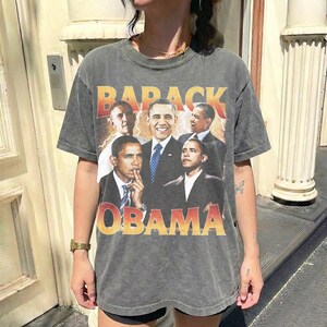 May include: A gray t-shirt featuring a collage of Barack Obama in various poses. The shirt has the words "BARACK OBAMA" in a distressed orange and yellow font. The shirt is worn by a person.