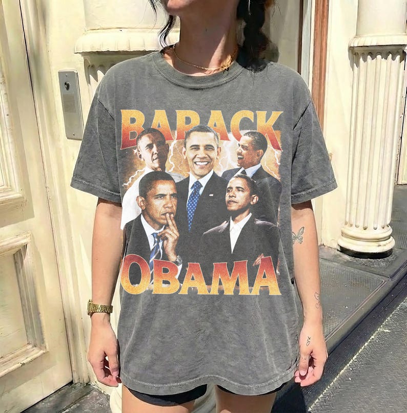 May include: A gray t-shirt featuring a collage of Barack Obama in various poses. The shirt has the words "BARACK OBAMA" in a distressed orange and yellow font. The shirt is worn by a person.