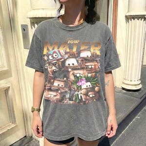 Puede incluir: Una camiseta gris de manga corta con un gr&aacute;fico de los personajes de la pel&iacute;cula "Cars". El dise&ntilde;o incluye el texto "Tow Mater" en letras doradas sobre la imagen de los personajes. La camiseta tiene un aspecto vintage y desgastado.