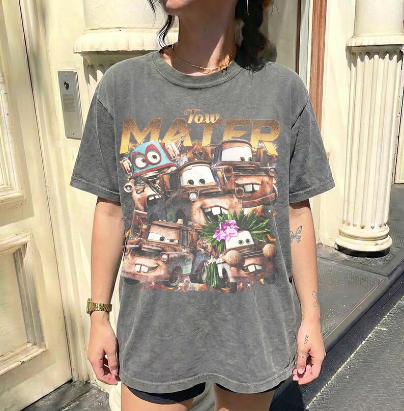Puede incluir: Una camiseta gris de manga corta con un gr&aacute;fico de los personajes de la pel&iacute;cula "Cars". El dise&ntilde;o incluye el texto "Tow Mater" en letras doradas sobre la imagen de los personajes. La camiseta tiene un aspecto vintage y desgastado.