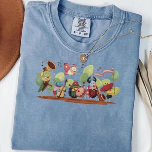 Puede incluir: Una camiseta azul de manga corta con un estampado gráfico de un grupo de insectos antropomórficos tocando instrumentos musicales. La camiseta tiene una etiqueta de la marca Comfort Colors en el cuello con el texto S-P-CH. Un collar con colgante dorado descansa sobre la tela.