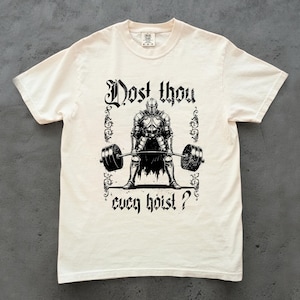 Könnte beinhalten: Cremefarbenes T-Shirt mit einer schwarzen Grafik eines Ritters, der eine Langhantel hebt. Der Text "Dost thou even hoist?" befindet sich über und unter der Ritterillustration. Das Shirt hat einen Rundhalsausschnitt und kurze Ärmel.