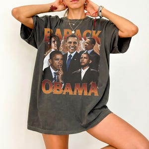 May include: Dark gray t-shirt featuring a graphic of Barack Obama in various poses. The word "BARACK" is printed above the images, and "OBAMA" is printed below in an orange gradient font. The shirt has short sleeves and a relaxed fit.