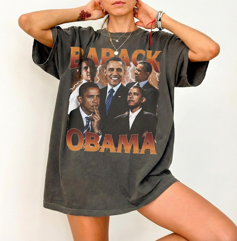 May include: Dark gray t-shirt featuring a graphic of Barack Obama in various poses. The word "BARACK" is printed above the images, and "OBAMA" is printed below in an orange gradient font. The shirt has short sleeves and a relaxed fit.