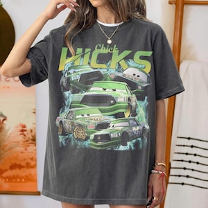 May include: Dark gray t-shirt featuring a graphic of Chick Hicks race cars. The design includes green cars with the number 86, the text "Chick Hicks" in yellow and green, and a blue and green background.