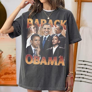 May include: Dark gray t-shirt featuring a collage of Barack Obama images with the text "BARACK OBAMA" in a gradient orange and red font. The shirt has a vintage, worn look.