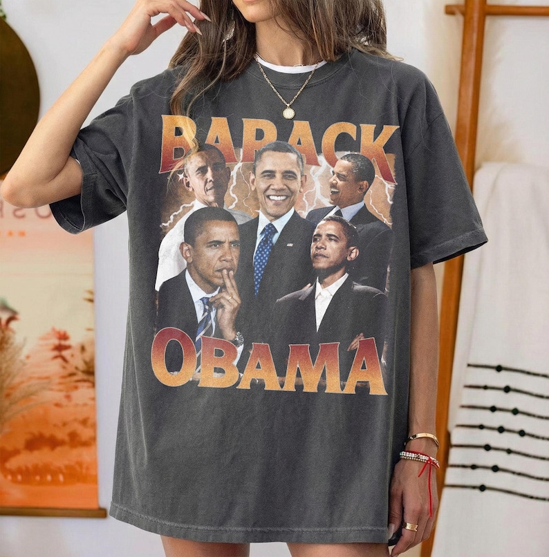 May include: Dark gray t-shirt featuring a collage of Barack Obama images with the text "BARACK OBAMA" in a gradient orange and red font. The shirt has a vintage, worn look.