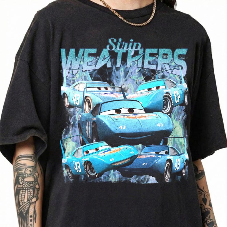 May include: Black t-shirt featuring a graphic of several blue cartoon race cars with the number 43. The text "Strip Weathers" is printed above the cars in a blue font. The cars are set against a blue and green background.