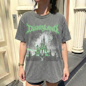 May include: Gray t-shirt with a distressed look, featuring the word "DISNEYLAND" in green, stylized lettering above a green-lit castle illustration with fireworks. The shirt has short sleeves and a crew neck.