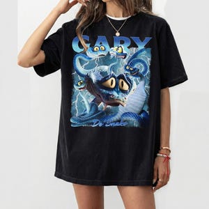 Pode incluir: T-shirt preto com uma ilustra&ccedil;&atilde;o de desenho animado de uma cobra azul com v&aacute;rias cabe&ccedil;as e o texto "Gary Dr. Snake" em azul. O gr&aacute;fico tem um visual vintage e desgastado. A camisa tem um ajuste folgado.