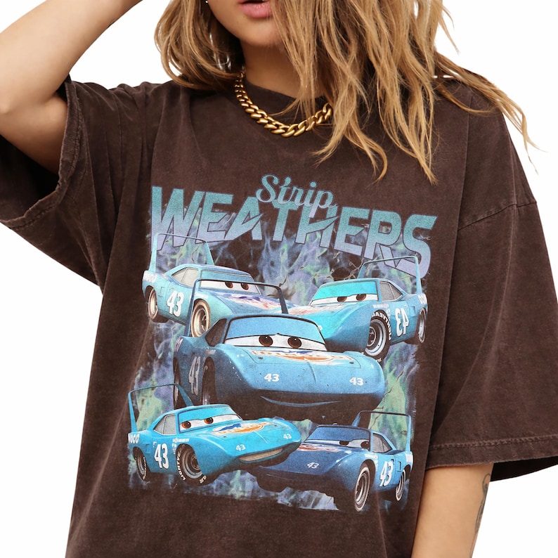 May include: Brown t-shirt featuring a graphic of blue race cars with the text "Strip Weathers". The cars have the number 43 on them. The shirt has a vintage, worn look.