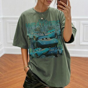 May include: Olive green oversized t-shirt featuring a graphic of blue race cars with the number 43. The text "Strip Weathers" is printed above the cars. The shirt has short sleeves and a relaxed fit.