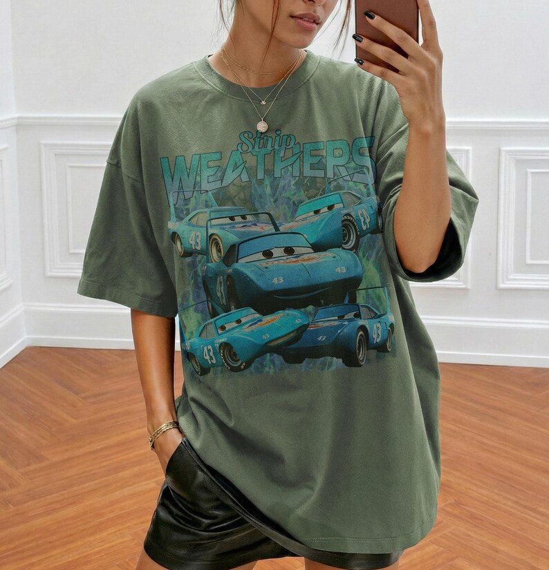 May include: Olive green oversized t-shirt featuring a graphic of blue race cars with the number 43. The text "Strip Weathers" is printed above the cars. The shirt has short sleeves and a relaxed fit.