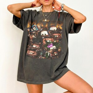 Puede incluir: Camiseta gris oscuro de gran tama&ntilde;o con un gr&aacute;fico del personaje Tow Mater de la pel&iacute;cula Cars. El gr&aacute;fico incluye el texto "Tow Mater" en letras doradas y varias im&aacute;genes del personaje. La camiseta tiene mangas cortas y un corte holgado.
