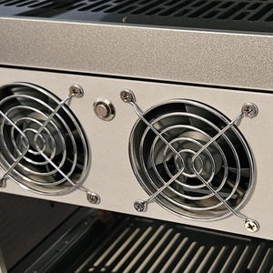 May include: A silver computer case component featuring two circular cooling fans protected by chrome wire grilles. The fans are mounted on a light gray panel secured with visible screws, positioned below a horizontal silver frame and above a vented base.