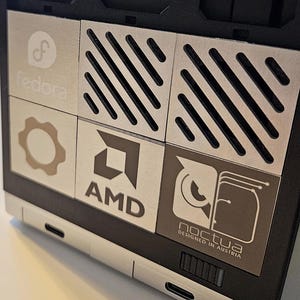 Może przedstawiać: Zbliżenie czarnego i srebrnego elementu komputera z kilkoma metalowymi panelami. Panele zawierają logo Fedora, AMD i Noctua, a także symbol koła zębatego i ukośne linie. Widoczny jest tekst "DESIGNED IN AUSTRIA".