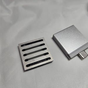 Puede incluir: Dos componentes electrónicos plateados. Uno es un pequeño bloque rectangular con un puerto USB-C. El otro es una pequeña placa rectangular con cinco ranuras negras paralelas.