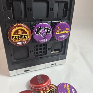 May include: A close-up of a black and silver computer case with several soda bottle caps attached. The caps are red, purple, and brown, featuring the brands "Coca-Cola", "Sunset Sarsaparilla", and "Quantum Jones".
