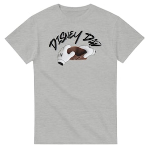 May include: A light gray t-shirt featuring the text "DISNEY DAD" in a handwritten style. The design includes a brown wallet held by white gloved hands, a nod to a famous cartoon character.