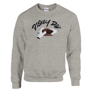 May include: A gray sweatshirt with the text "DISNEY DAD" in a black script font. The design includes cartoon hands holding a brown object. The sweatshirt is made of a soft material.