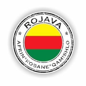 May include: A circular sticker featuring the word "ROJAVA" at the top and "AFRIN*KOBANE*QAMISHL" around the bottom. The centre displays a flag with yellow, red, and green horizontal stripes. The sticker has a distressed, vintage appearance.