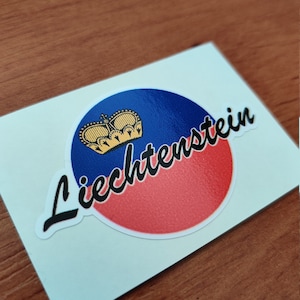 Peut inclure: Un autocollant blanc avec un cercle rouge et bleu représentant une couronne dorée et le mot "Liechtenstein" en écriture cursive noire.