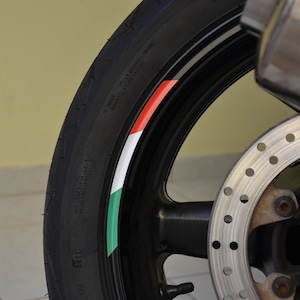 May include: Close-up of a motorcycle tire with a black, white, green, and red stripe design. The tire is mounted on a black rim with a silver brake disc.
