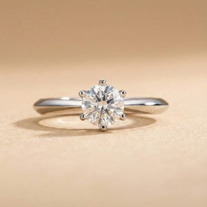 Może przedstawiać: Pierścionek zaręczynowy z brylantem o okrągłym szlifie, osadzonym w sześcioramiennej oprawie. Obrączka wykonana jest z polerowanego srebrnego metalu. Pierścionek jest prezentowany na neutralnym tle.