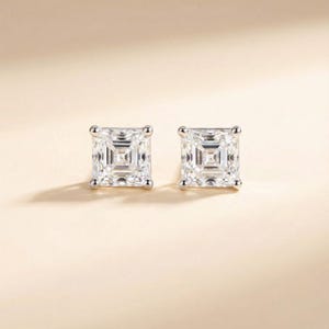 Op de afbeelding: Een paar vierkant geslepen diamant-oorbellen. De oorbellen zijn gezet in een zilverkleurig metaal en worden gepresenteerd tegen een lichtbeige achtergrond. De diamanten hebben een briljante, facetrijke uitstraling en reflecteren licht.