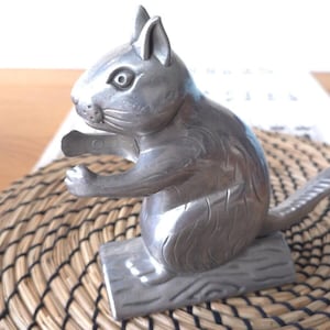 May include: A silver-toned metal nutcracker shaped like a squirrel sitting on a log. The squirrel has detailed fur textures, pointed ears, and a long tail extending backward. The functional mechanism is integrated into the squirrel's front paws.