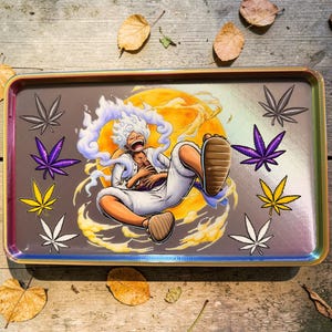 Könnte beinhalten: Rechteckige Rolling Tray mit farbigem Rand. Die Tray zeigt eine Cartoonfigur mit weißen Haaren und Kleidung, vor einem gelb-orangen Hintergrund. Marihuanablätter in verschiedenen Farben sind ebenfalls auf der Tray abgebildet.
