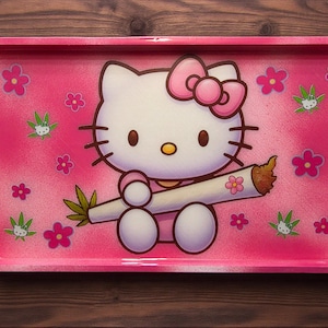 May include: Pink rectangular rolling tray featuring a cartoon cat character with a pink bow, holding a lit joint. The tray is decorated with pink flowers and cannabis leaf designs.
